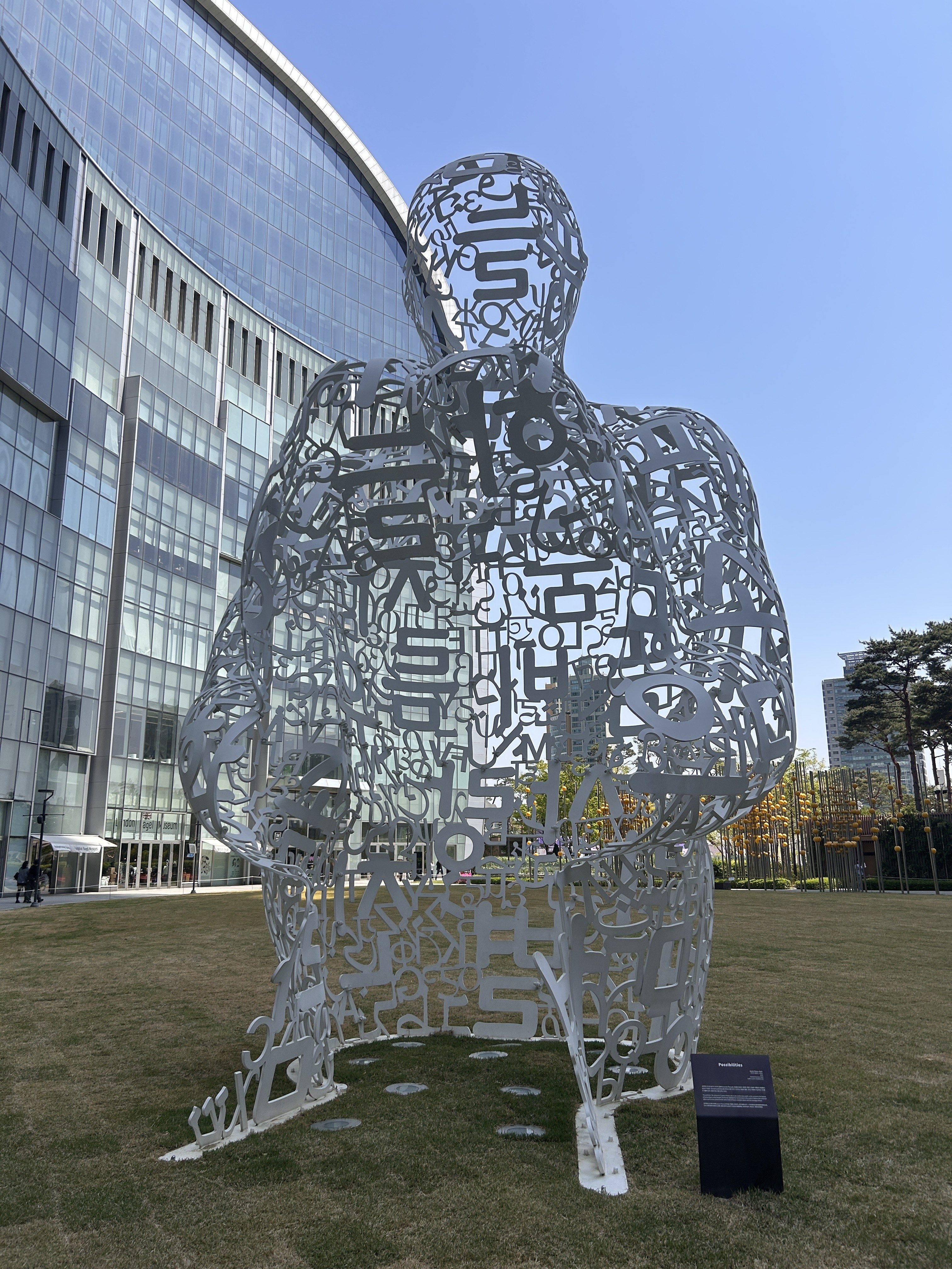 A large, white sculpture resembling a human figure, made up of various letters and symbols, stands on a grassy area in front of a modern glass building