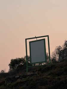 A large green frame stands on a grassy slope, with a blank white panel inside it. A small bird perches on top of the frame, while the background features a soft, hazy sky at dusk.