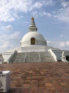 A large white stupa with a golden Buddha statue in a niche, surrounded by stone steps leading up to it. The stupa is topped with a series of rings and a small golden structure. Fluffy clouds are visible in the sky above.