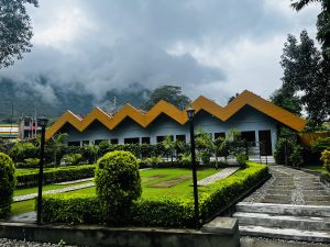 A row of buildings with yellow zigzag roofs is surrounded by a well-maintained garden and walkway, set against a backdrop of misty mountains and a cloudy sky.