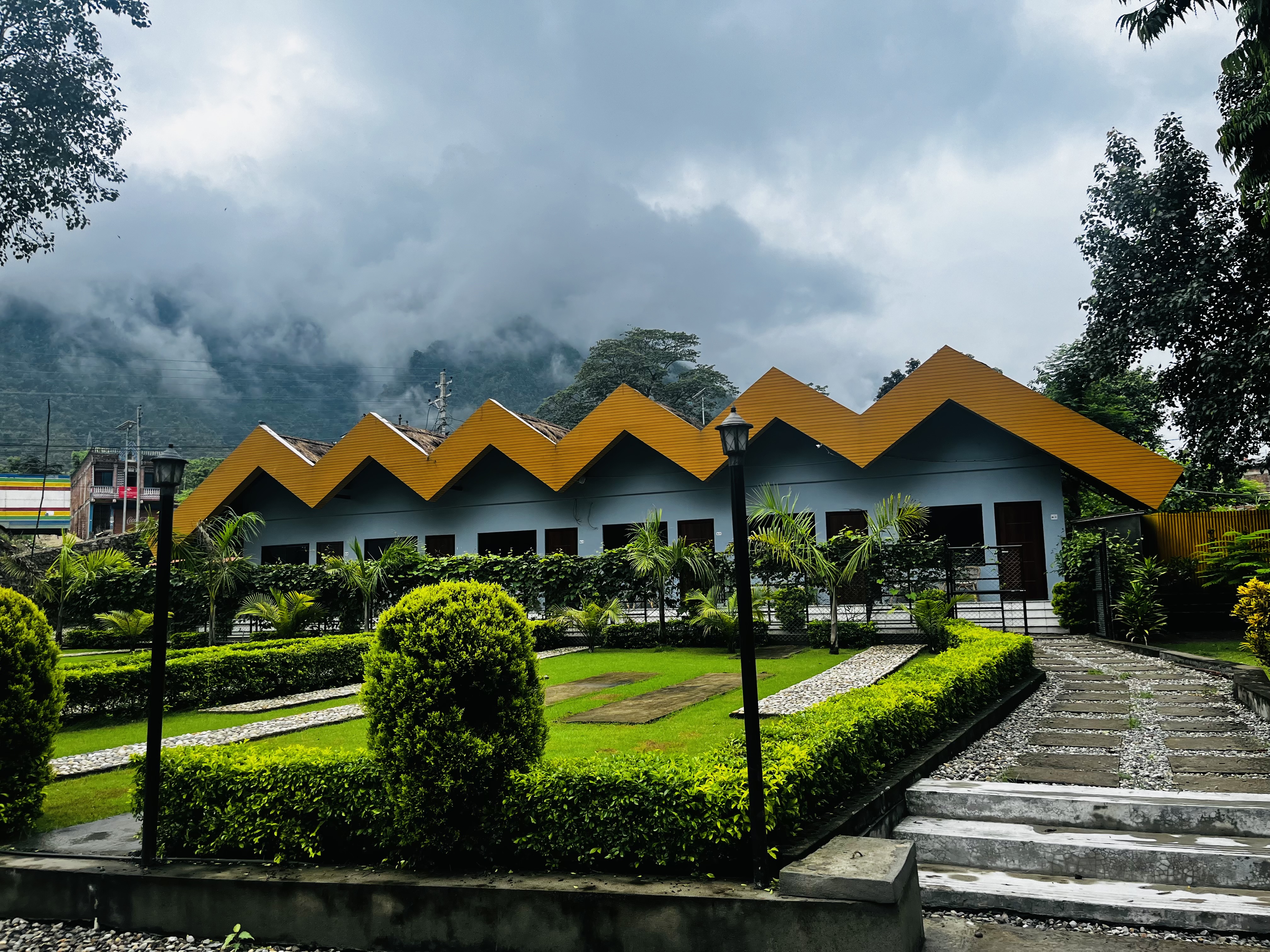 A row of buildings with yellow zigzag roofs is surrounded by a well-maintained garden and walkway, set against a backdrop of misty mountains and a cloudy sky.