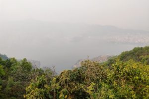 A panoramic view of a large lake surrounded by lush green trees and vegetation, with hills in the background shrouded in haze.