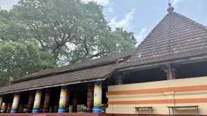 A traditional building with a tiled roof and a triangular gable, surrounded by lush green trees under a partly cloudy sky. The structure features a row of colorful pillars painted in red, yellow, and white, adorned with intricate designs.
