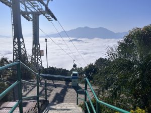 A scenic view from a hilltop featuring a cable car against a backdrop of mountains and a sea of clouds. In the foreground, there are stairs with green railings leading downwards, surrounded by lush greenery and trees. A clear blue sky completes the serene landscape.
