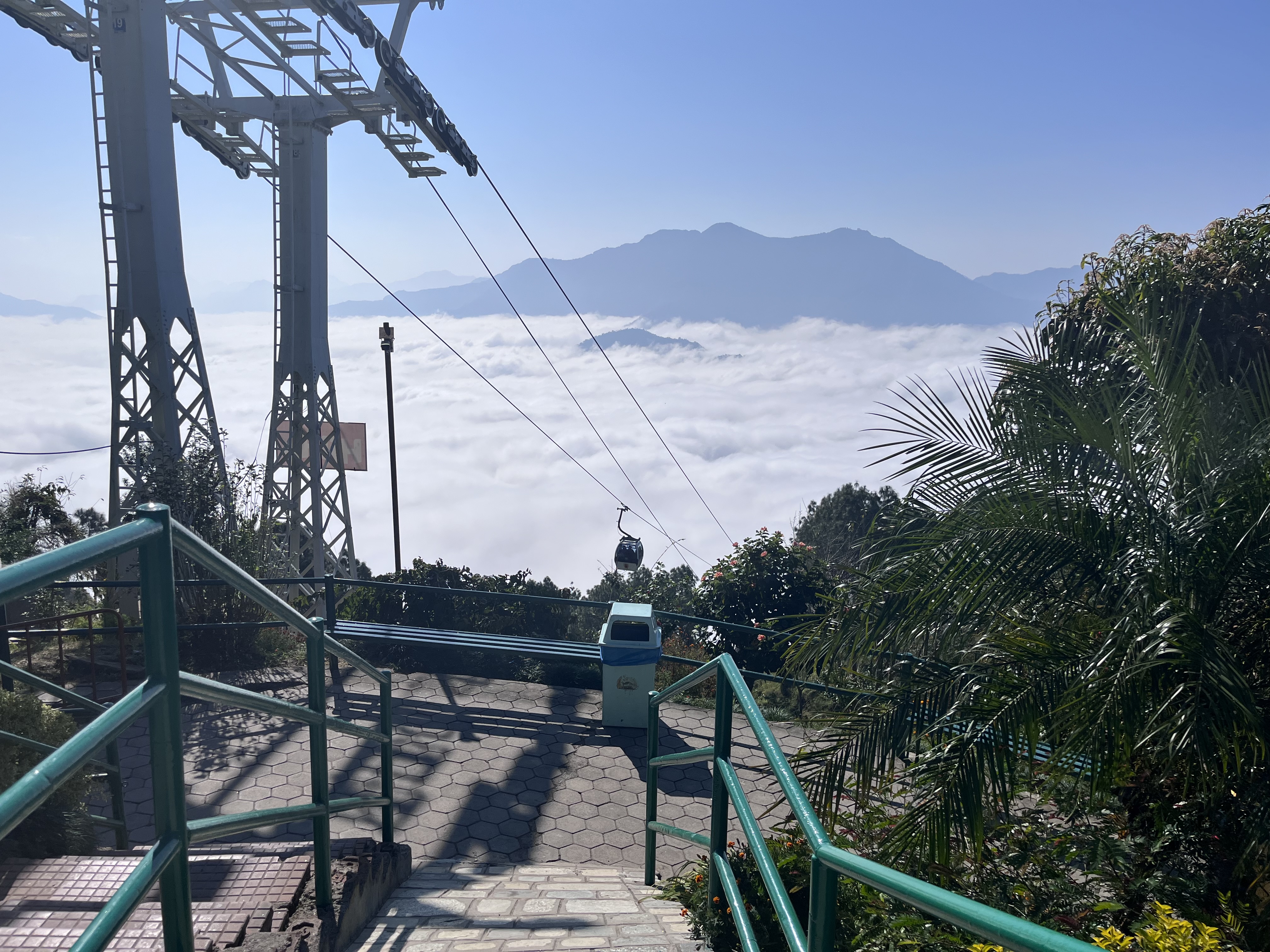 A scenic view from a hilltop featuring a cable car against a backdrop of mountains and a sea of clouds. In the foreground, there are stairs with green railings leading downwards, surrounded by lush greenery and trees. A clear blue sky completes the serene landscape.