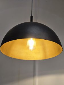 A pendant light with a black, dome-shaped exterior and a golden interior. The light bulb is visible and lit, providing a warm glow that reflects off the golden interior of the lampshade. Spotted it at a restaurant in Kozhikode, Kerala. #WPNP2025
