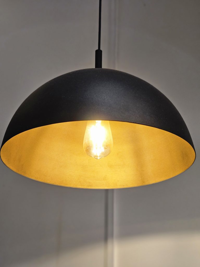 A pendant light with a black, dome-shaped exterior and a golden interior. The light bulb is visible and lit, providing a warm glow that reflects off the golden interior of the lampshade. Spotted it at a restaurant in Kozhikode, Kerala. #WPNP2025