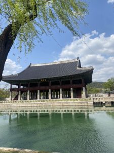 A traditional East Asian pavilion stands by a serene body of water, reflecting the structure in its greenish surface.