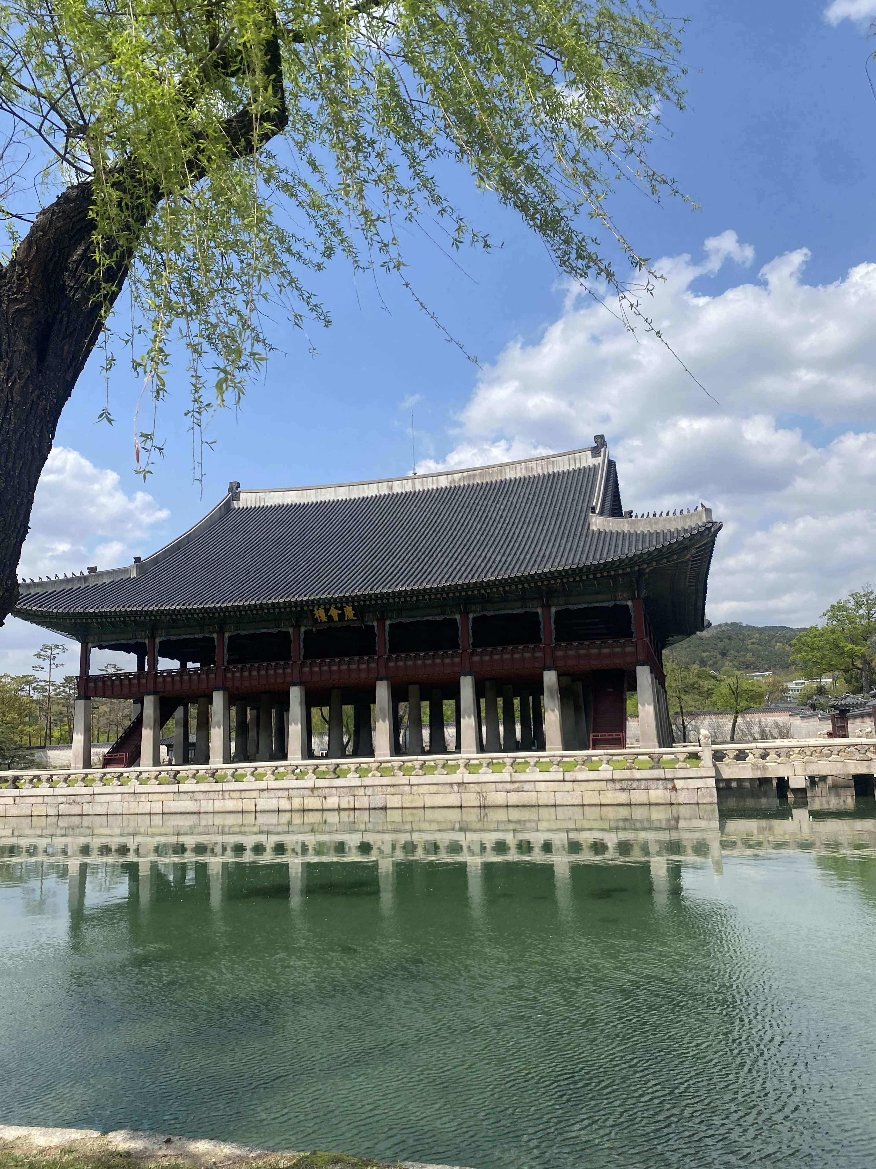 A traditional East Asian pavilion stands by a serene body of water, reflecting the structure in its greenish surface.