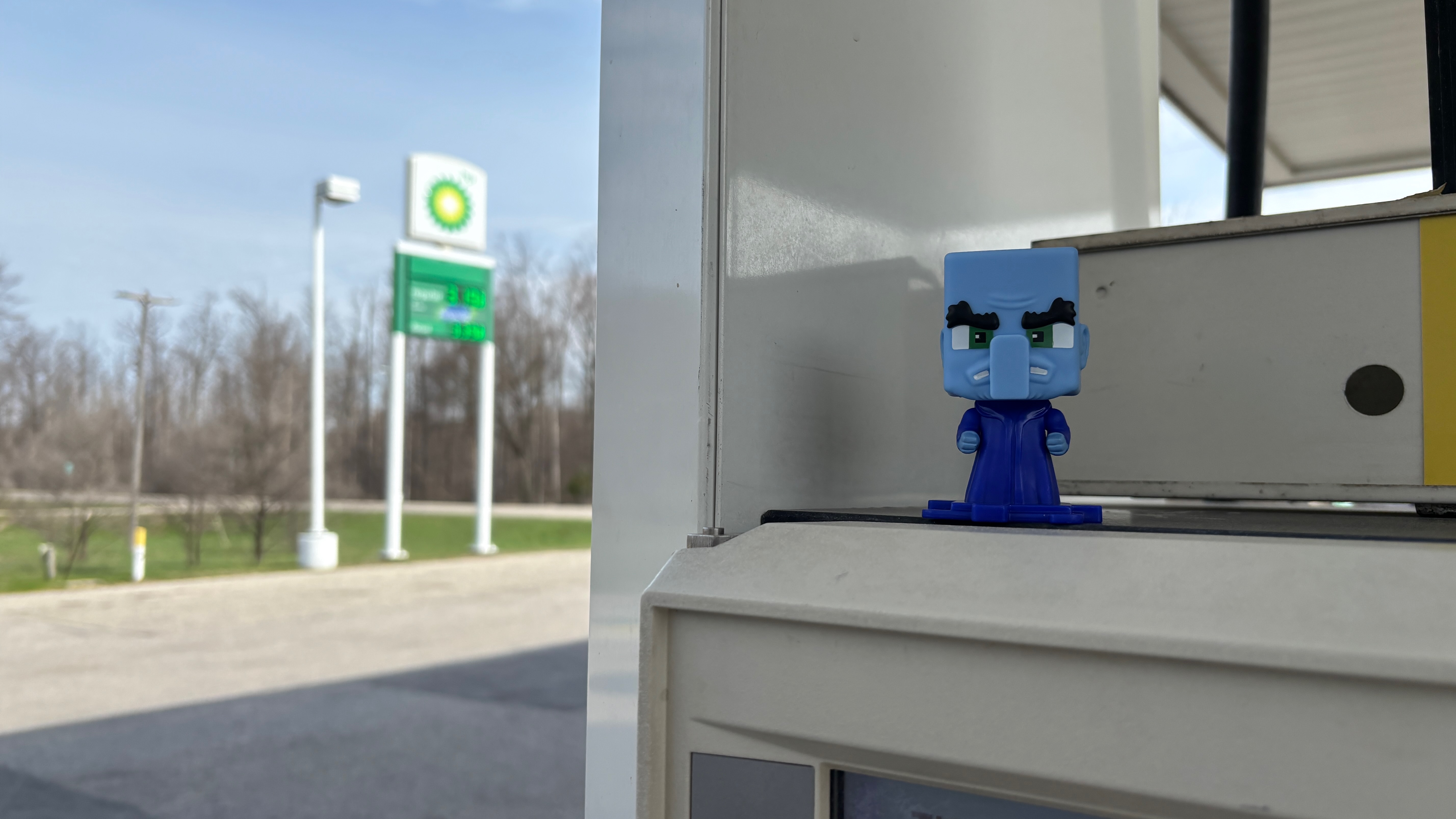 A small toy cartoon character sits atop a gas pump. The gas station sign is blurry in the background.
