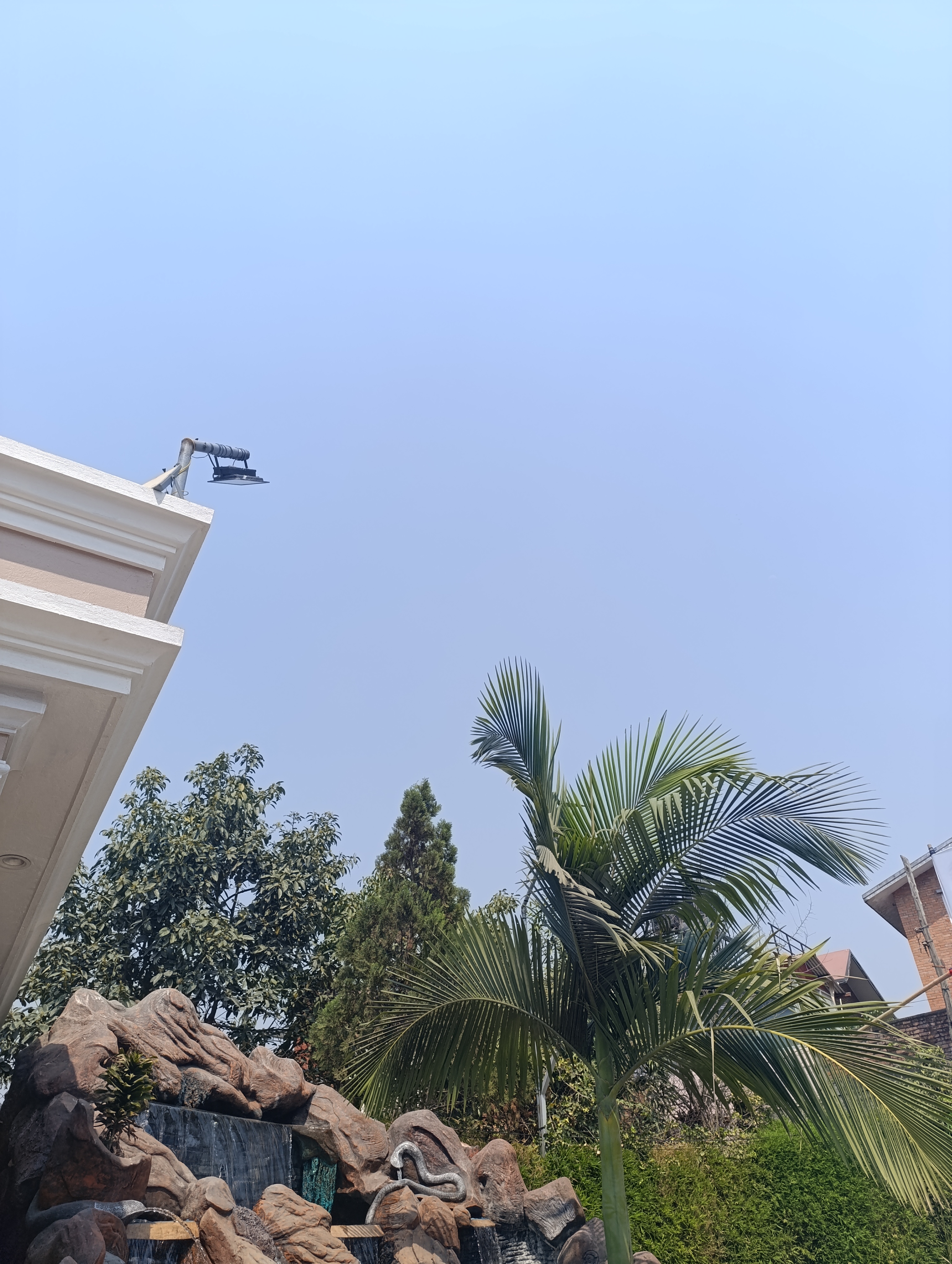 A clear sky with a portion of a building roof in the top left, featuring a mounted light fixture. Below, a landscaped garden with a rock waterfall, tall palm tree, and a variety of other lush green trees and plant life.