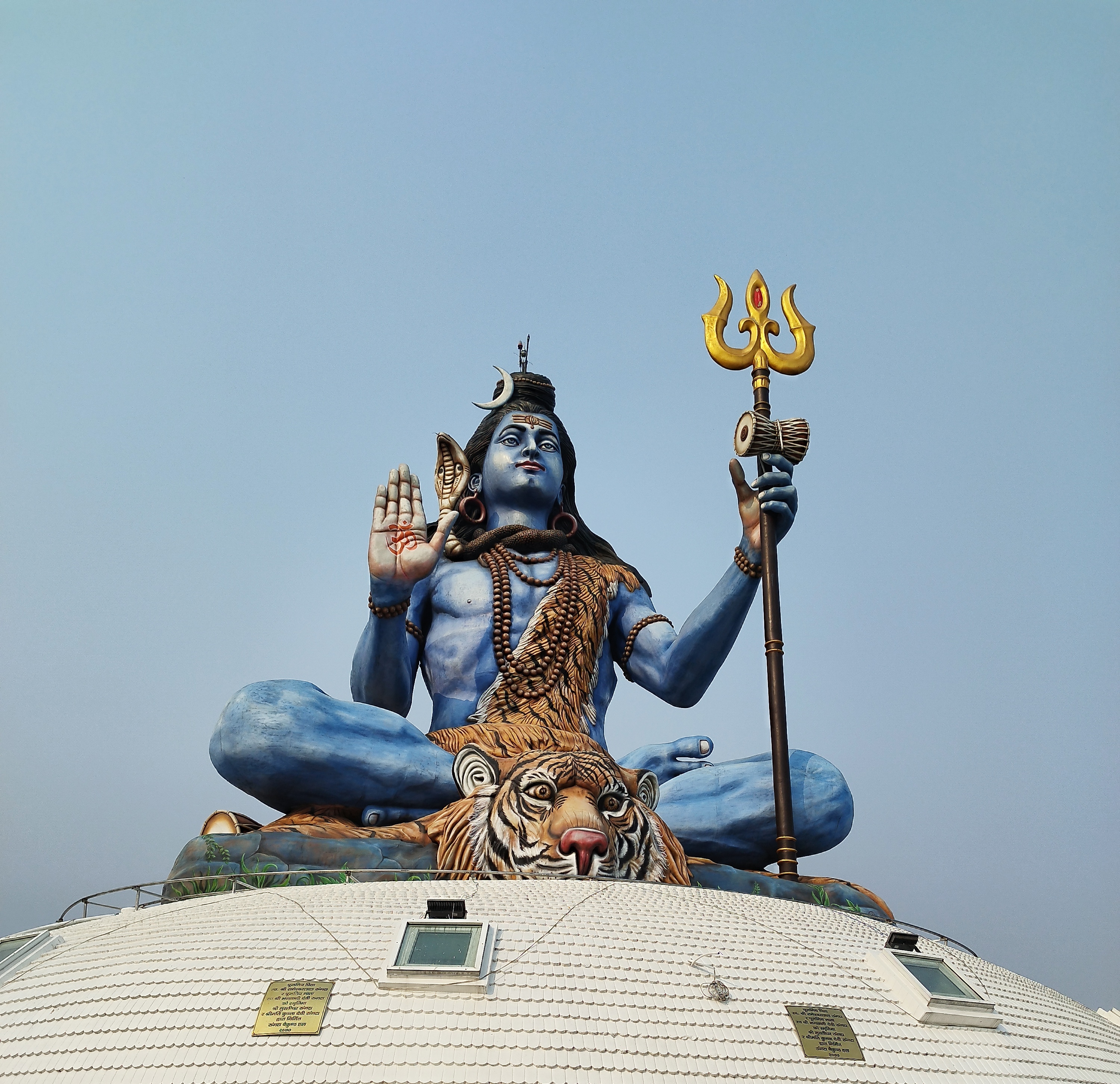 A Pumdikot Shiva statue of a blue-skinned deity sitting cross-legged atop a dome. The figure has long hair, wears a tiger skin, holds a trident, and features a crescent moon on its head. The background is a clear blue sky.