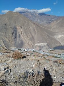 A mountainous landscape with a rugged terrain and sparse vegetation in the foreground. In the distance, there is a small village with buildings and fields, surrounded by towering mountains partly covered in clouds. The sky is clear and blue.