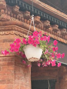 A white hanging planter filled with vibrant pink petunia flowers is suspended from a rustic brick wall adorned with traditional carvings.