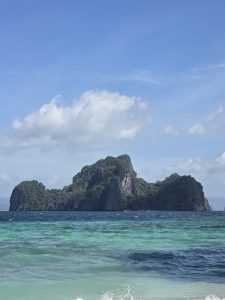 A stunning view from El Nido, Palawan, featuring turquoise waters, gentle waves, and a towering limestone island covered in lush greenery. The bright blue sky with scattered clouds adds to the serene, tropical beauty of this paradise.