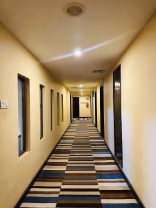 A long, well-lit hallway with beige walls, featuring a striped carpet. 