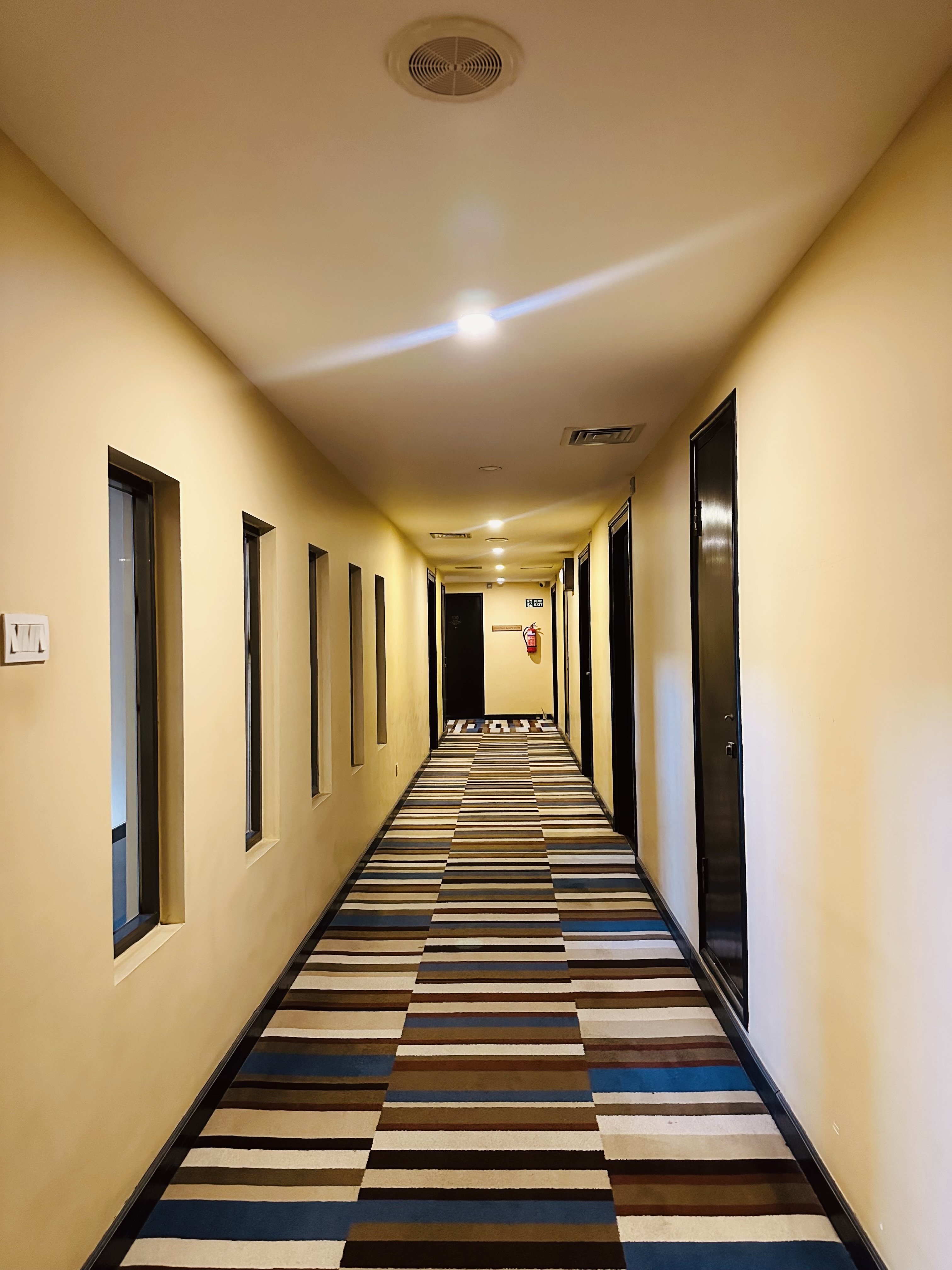A long, well-lit hallway with beige walls, featuring a striped carpet.