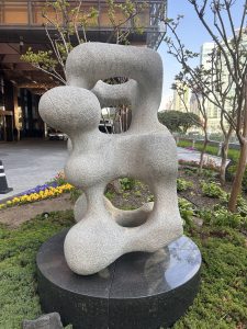 A large abstract stone sculpture featuring smooth, rounded forms and hollow openings. The sculpture is displayed on a dark circular pedestal amidst greenery and colorful flowers, with a modern building in the background.
