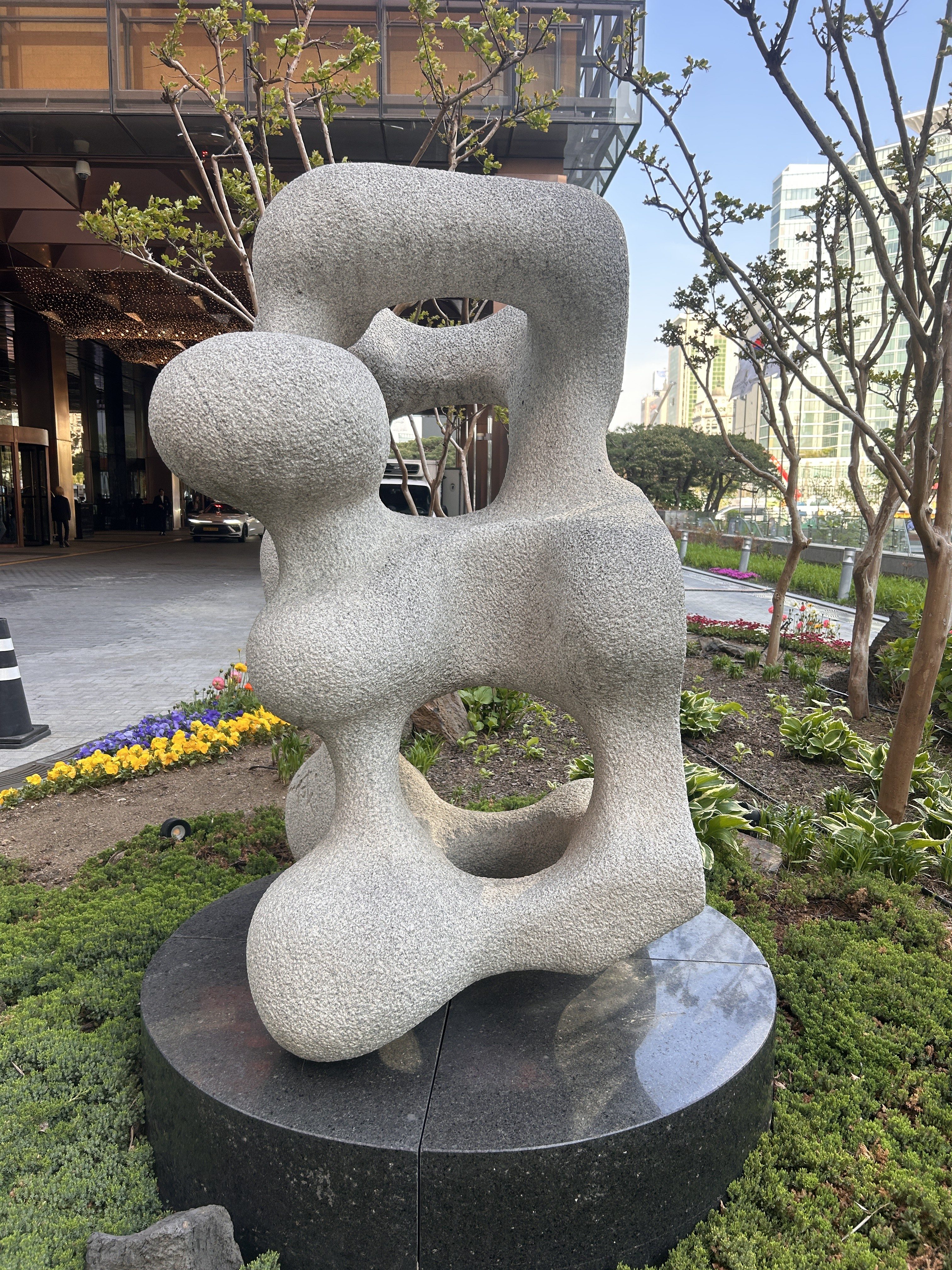 A large abstract stone sculpture featuring smooth, rounded forms and hollow openings. The sculpture is displayed on a dark circular pedestal amidst greenery and colorful flowers, with a modern building in the background.