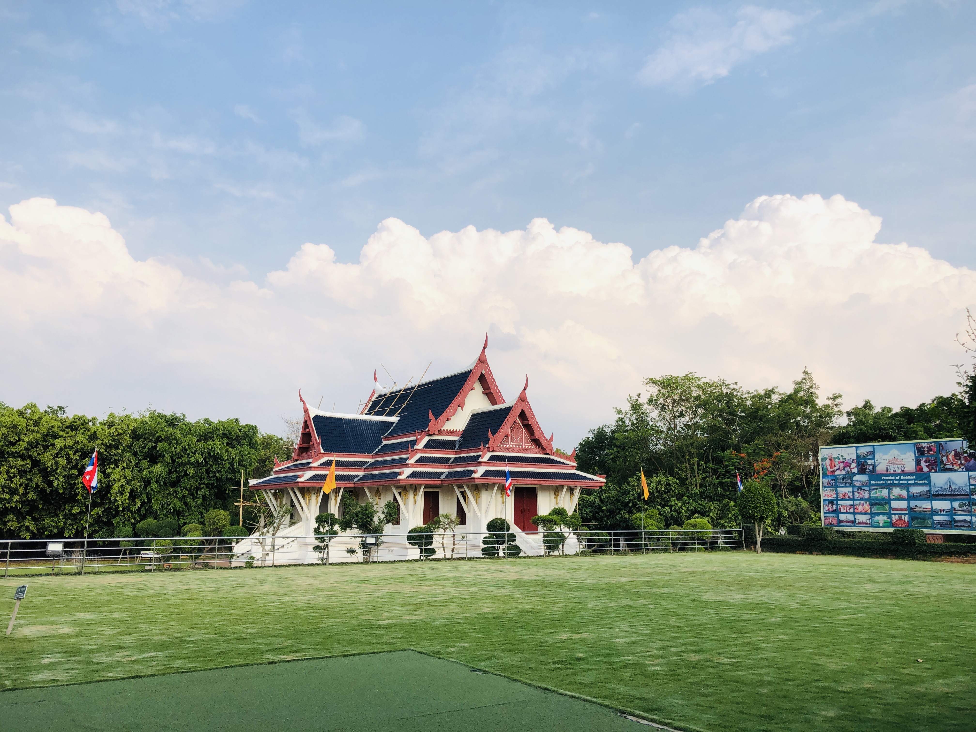 A beautifully ornate Thai-style temple sits peacefully in a lush green park, surrounded by national flags and vibrant billboards under a bright, cloud-filled sky.