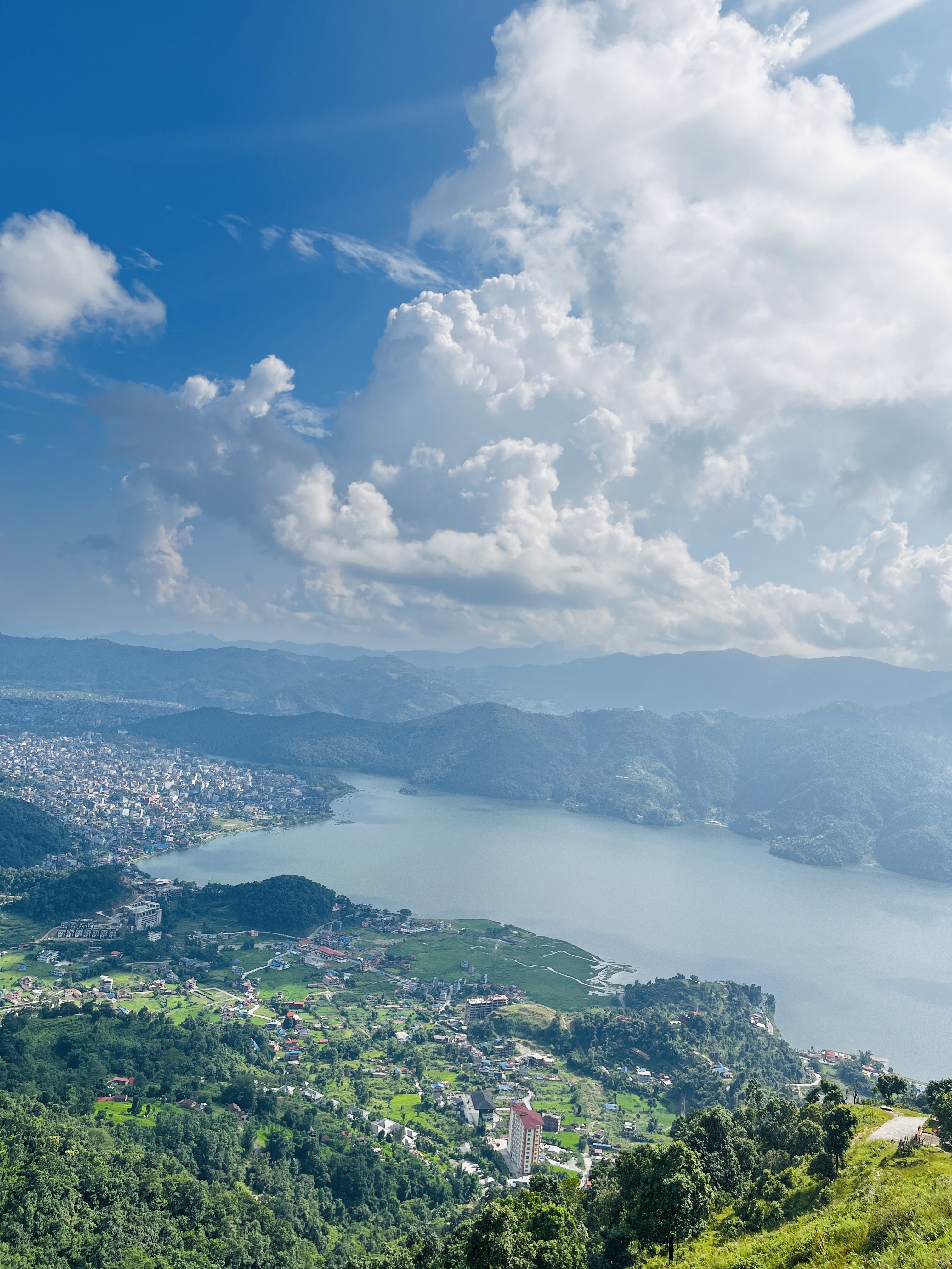 A scenic view of a large lake surrounded by lush green hills and a cityscape. The sky is filled with fluffy white clouds and has a bright blue hue.