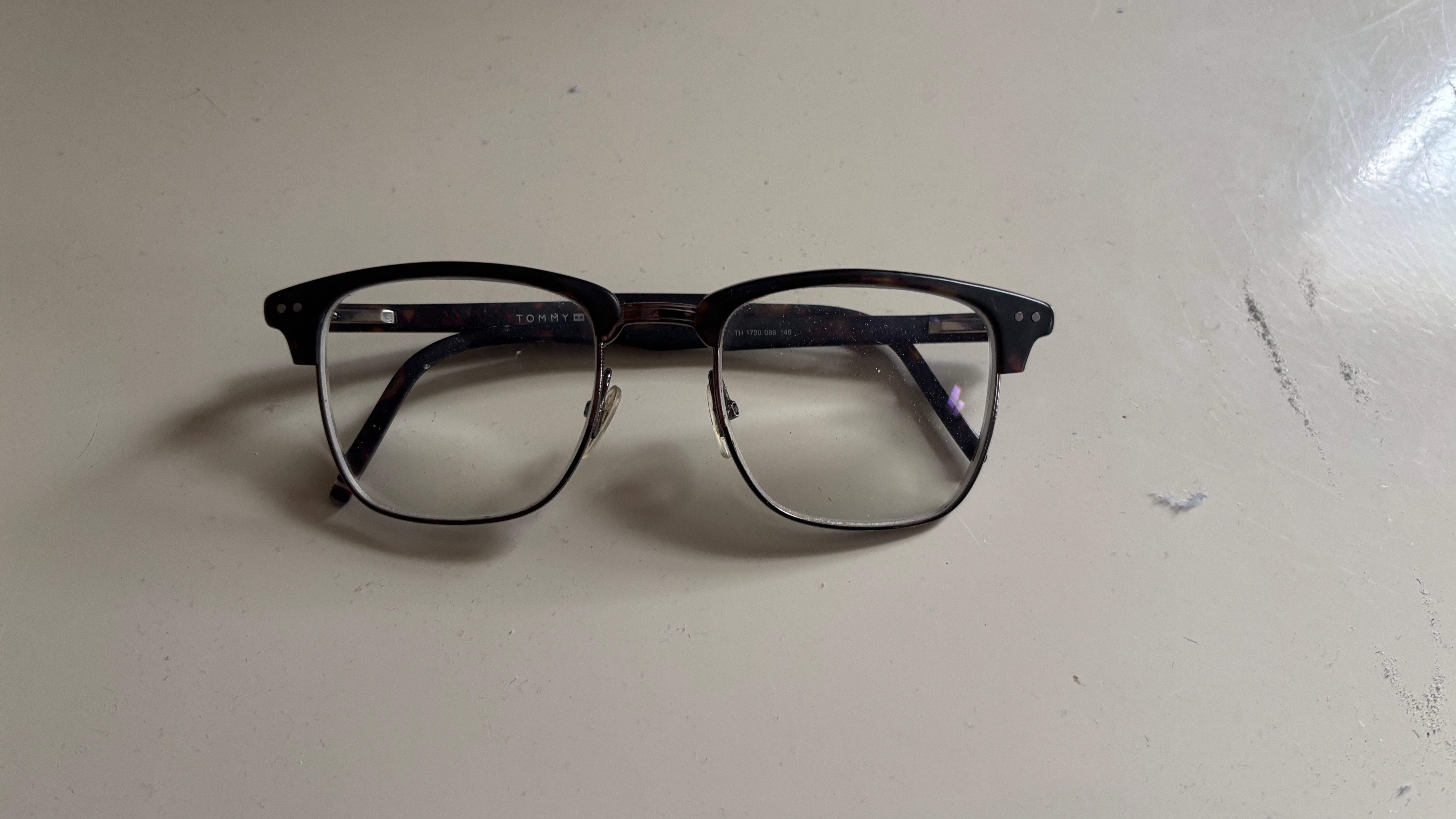 A pair of glasses is lying on a table. They have dark rims on the top and metal on the bottom. Light shines from the top left, making a shadow underneath.