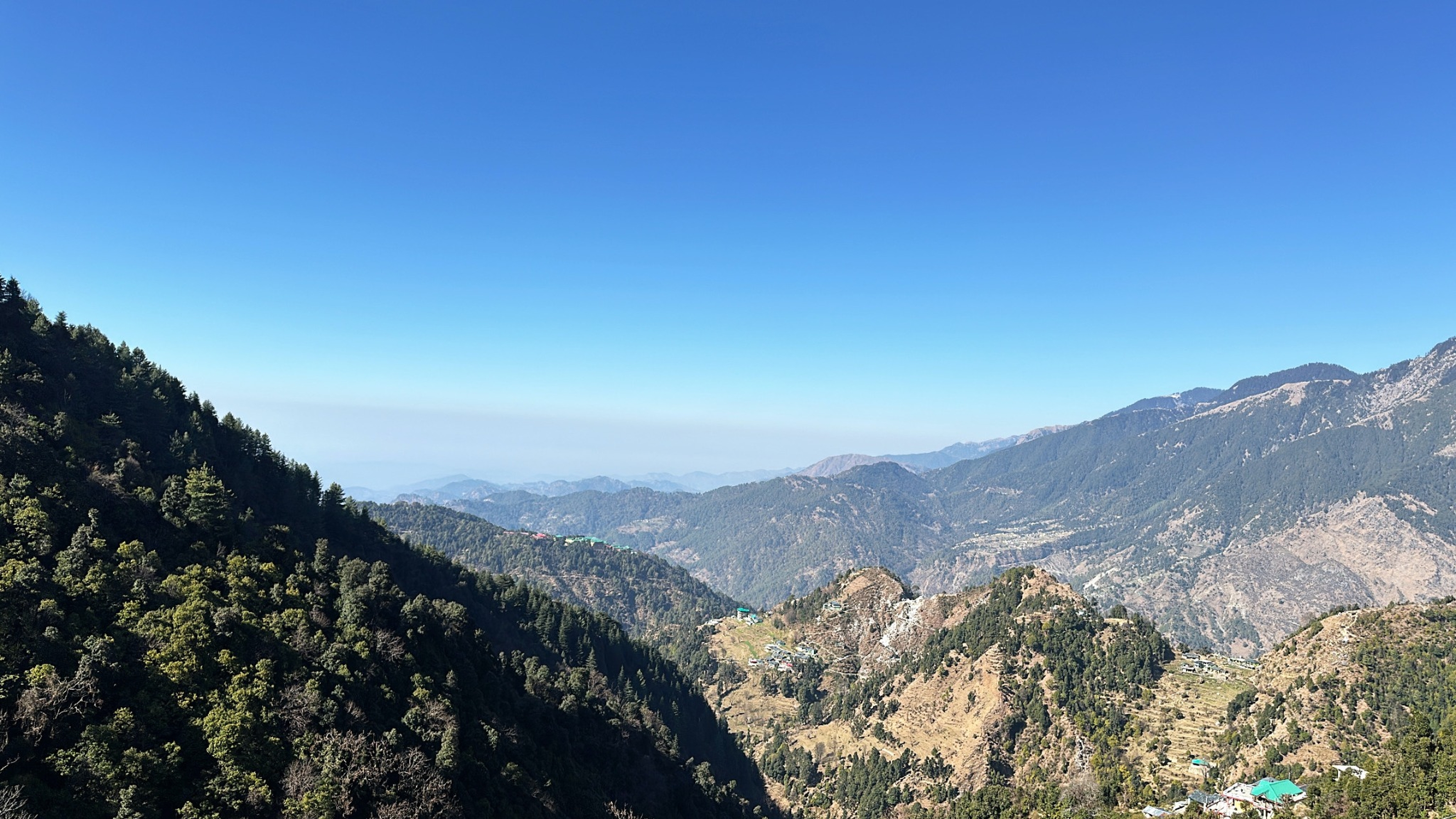 A panoramic view of a mountainous landscape with densely forested hills in the foreground and a clear blue sky above. The distant hills and valleys are visible under the bright daylight, showcasing a picturesque and serene natural setting.