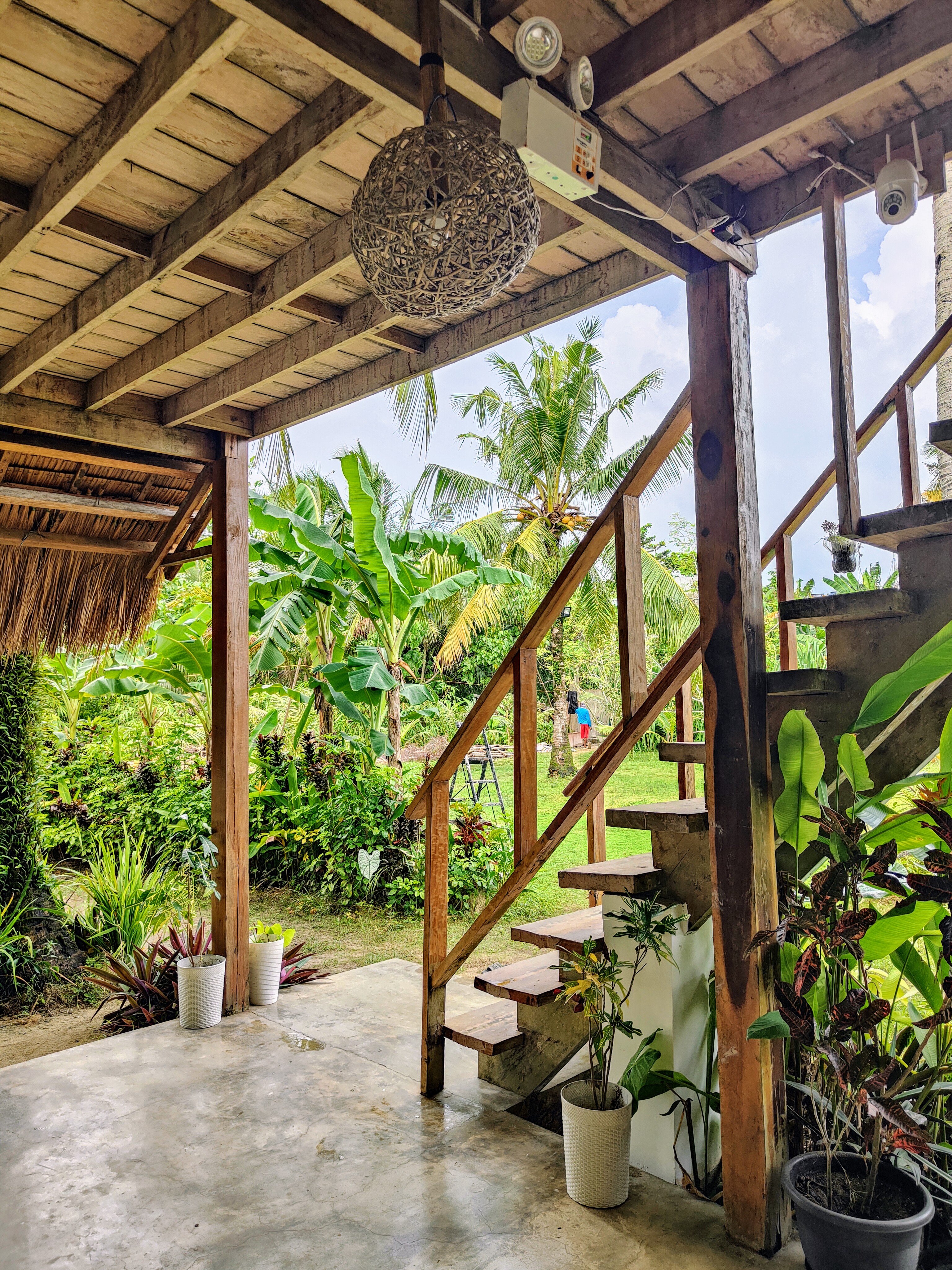A rustic porch in Siargao, Philippines, featuring wooden stairs, a woven pendant light, and potted plants. Surrounded by lush tropical greenery and banana trees, it captures the island’s serene and natural charm.