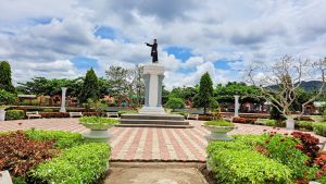 A well-maintained garden with colorful plants surrounds a large, elevated statue of a man in a black suit. The statue is set on a column pedestal at the center of a paved, red-and-beige patterned area.