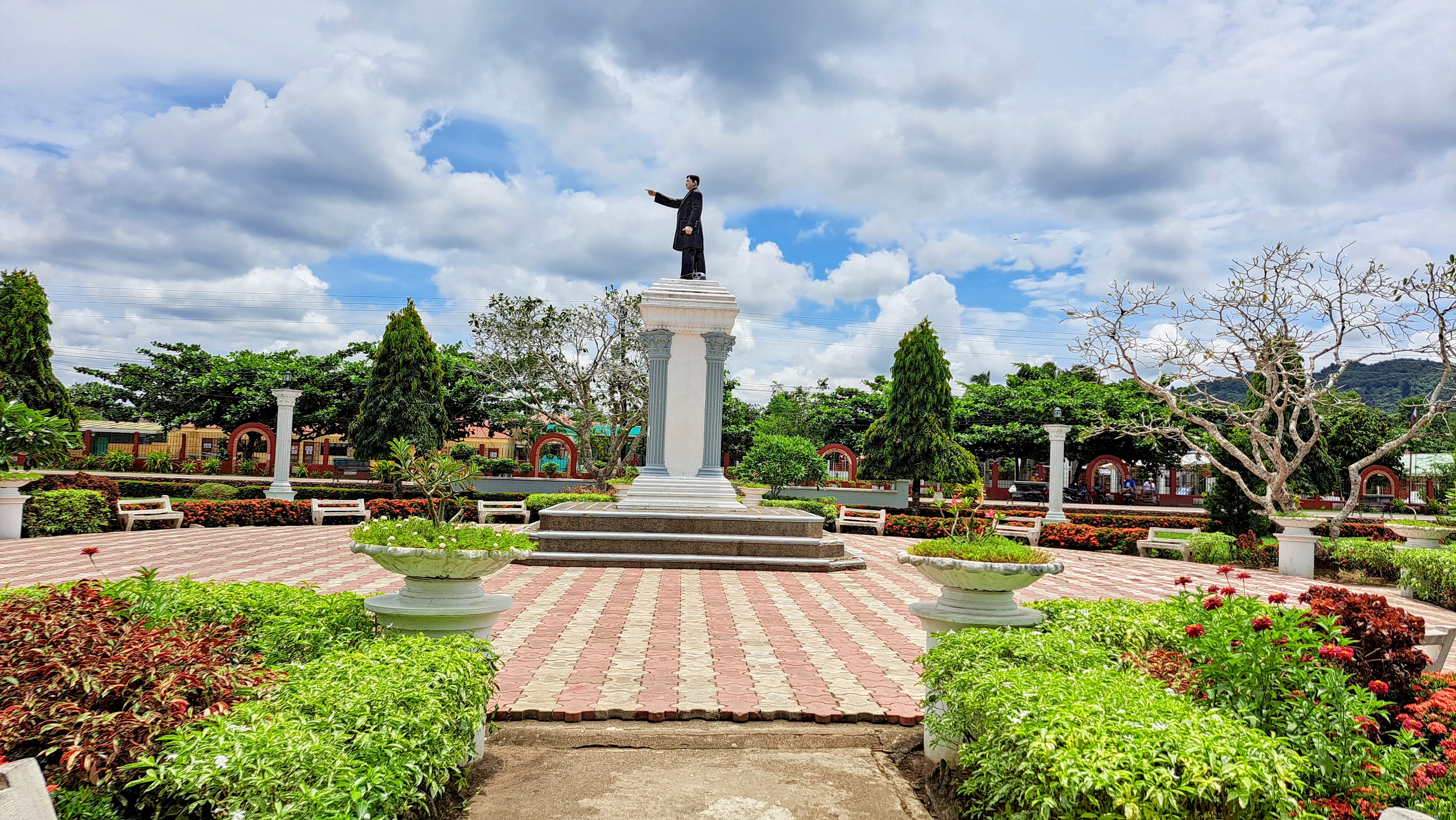A well-maintained garden with colorful plants surrounds a large, elevated statue of a man in a black suit. The statue is set on a column pedestal at the center of a paved, red-and-beige patterned area.