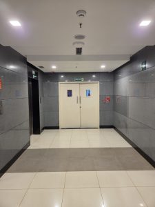 A hallway with glossy tiled floors and walls, featuring a set of double doors marked as a fire exit. On the left is an elevator with a closed door, and on the right, there are fire safety devices. The ceiling has recessed lighting and a smoke detector.