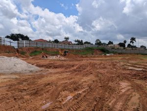 A construction site with a large, cleared dirt area under a partly cloudy sky. Several workers in orange vests are scattered across the site, engaging in various activities, including operating machinery. 