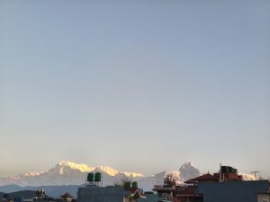 A scenic view of snow-capped mountains under a clear blue sky, with rooftops and water tanks in the foreground.