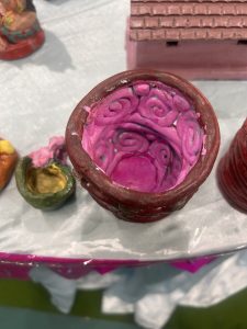 A close-up of a handcrafted clay pot with a red exterior and a pink interior featuring spiral designs. Other small clay objects and a model of a house are visible in the background.
