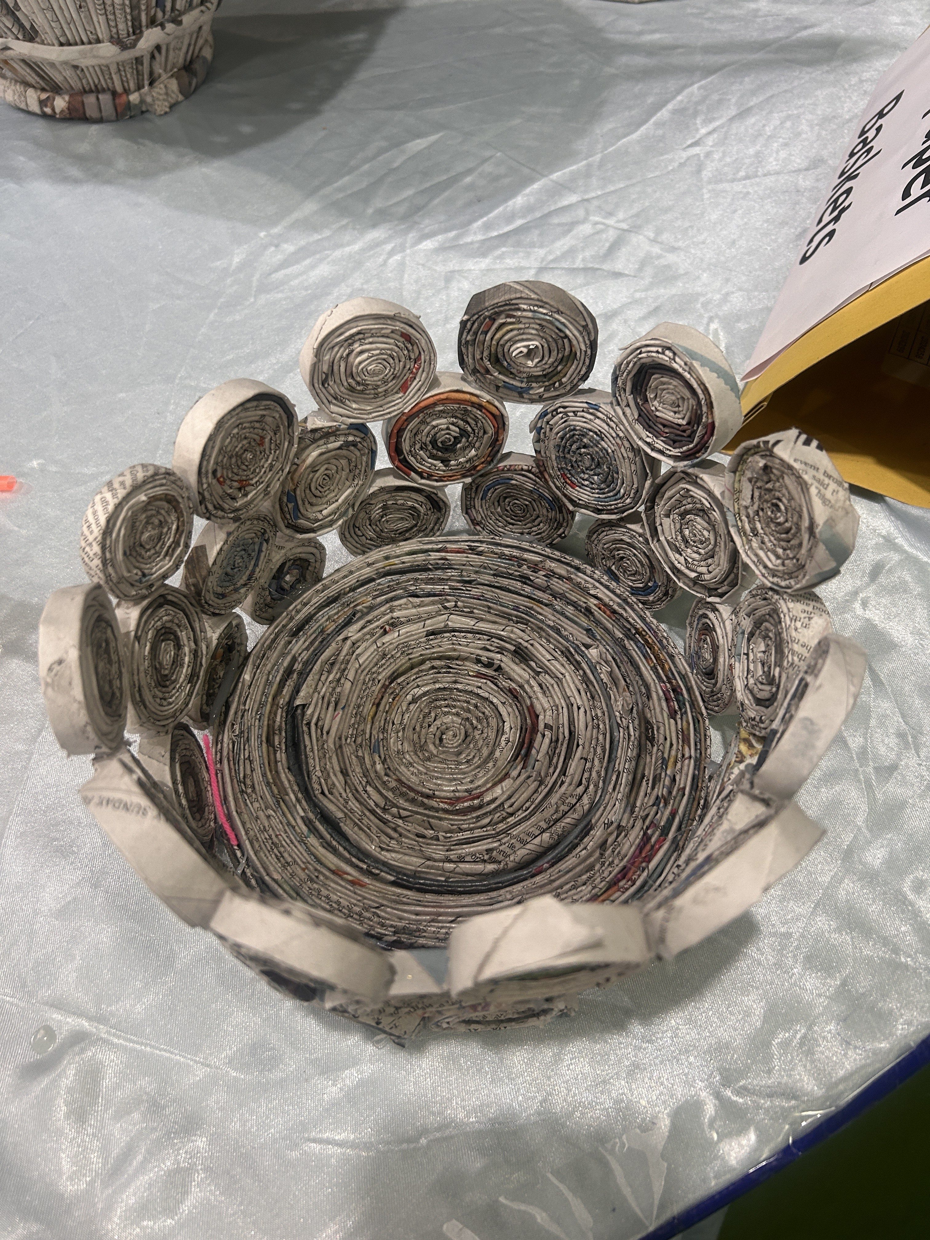 A handmade bowl crafted from rolled newspaper strips, arranged in a circular pattern, with a textured appearance