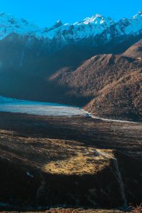 A breathtaking view of a mountainous landscape, featuring snow-capped peaks in the background, sunlit slopes in the foreground, and a lush valley with varying shades of brown and green.
