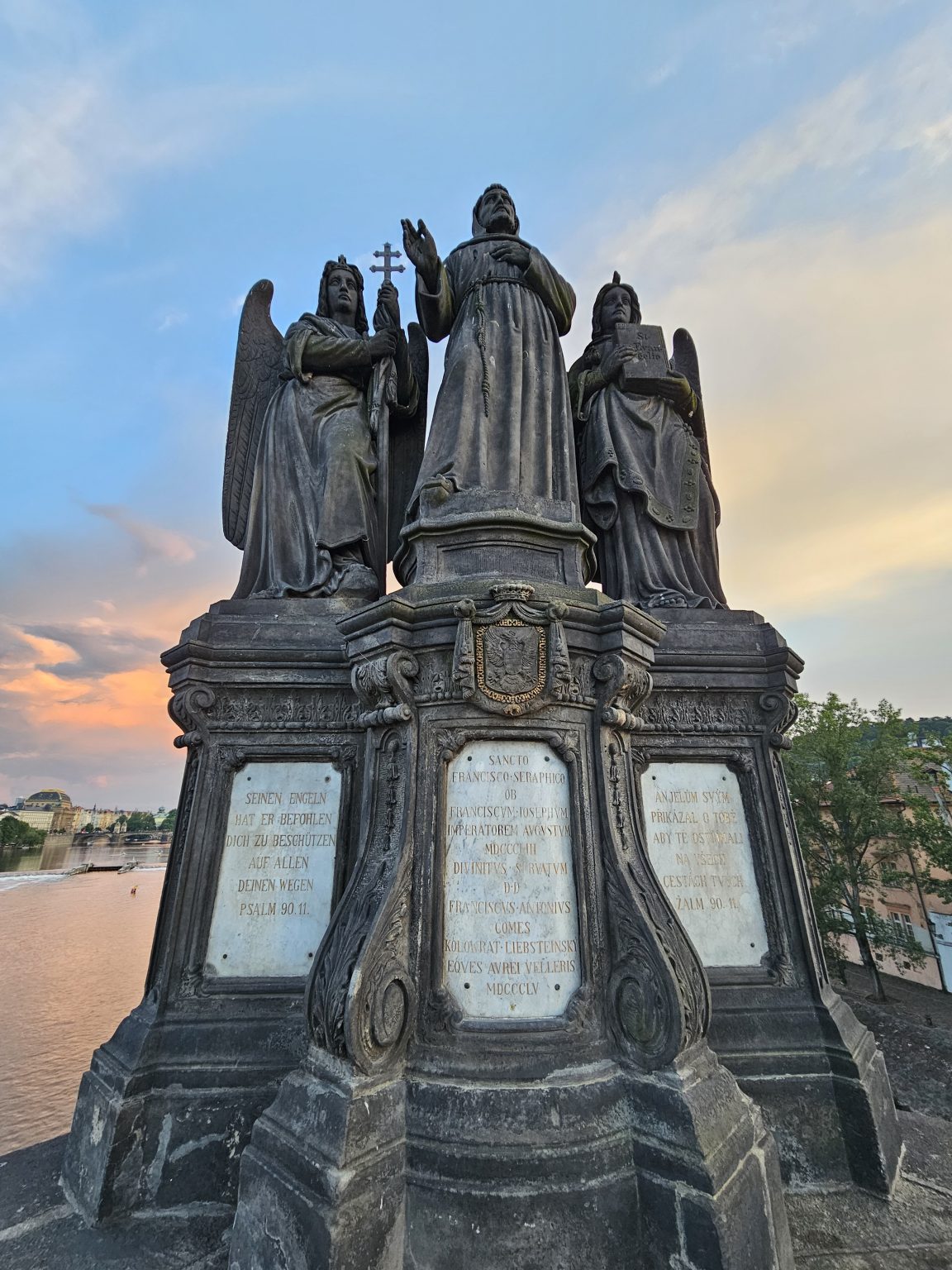 The image shows the Statue of Francis of Assisi on the Charles Bridge in Prague, featuring St. Francis flanked by angels and inscriptions on the base. The statue is made of dark stone and is set against a backdrop of the Vltava River and a colorful sky.