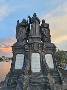 The image shows the Statue of Francis of Assisi on the Charles Bridge in Prague, featuring St. Francis flanked by angels and inscriptions on the base. The statue is made of dark stone and is set against a backdrop of the Vltava River and a colorful sky.
