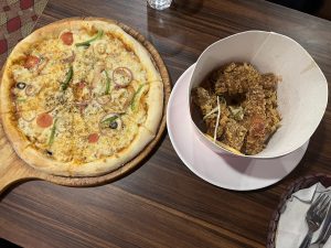 A freshly baked pizza topped with cheese, onions, bell peppers, tomatoes, and olives on a wooden board, next to a bucket of crispy fried chicken on a wooden table.