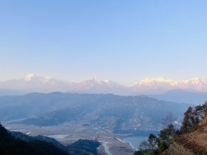 A panoramic view of a mountain range at dawn, with snow-capped peaks illuminated by the soft morning light. The foreground features rolling hills descending into a valley with a winding river and a small lake. Trees and patches of vegetation are visible on the slopes. The sky is clear with a hint of warm colors on the horizon.