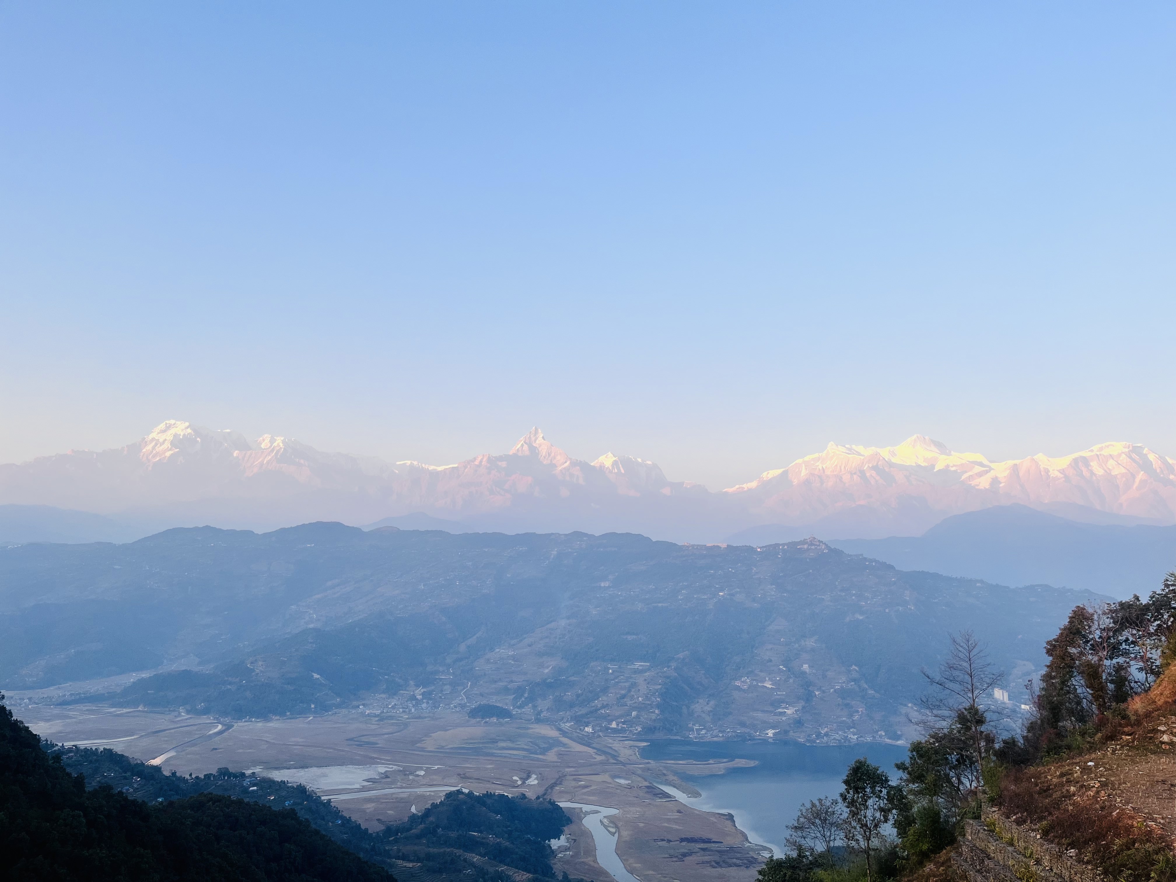 A panoramic view of a mountain range at dawn, with snow-capped peaks illuminated by the soft morning light. The foreground features rolling hills descending into a valley with a winding river and a small lake. Trees and patches of vegetation are visible on the slopes. The sky is clear with a hint of warm colors on the horizon.