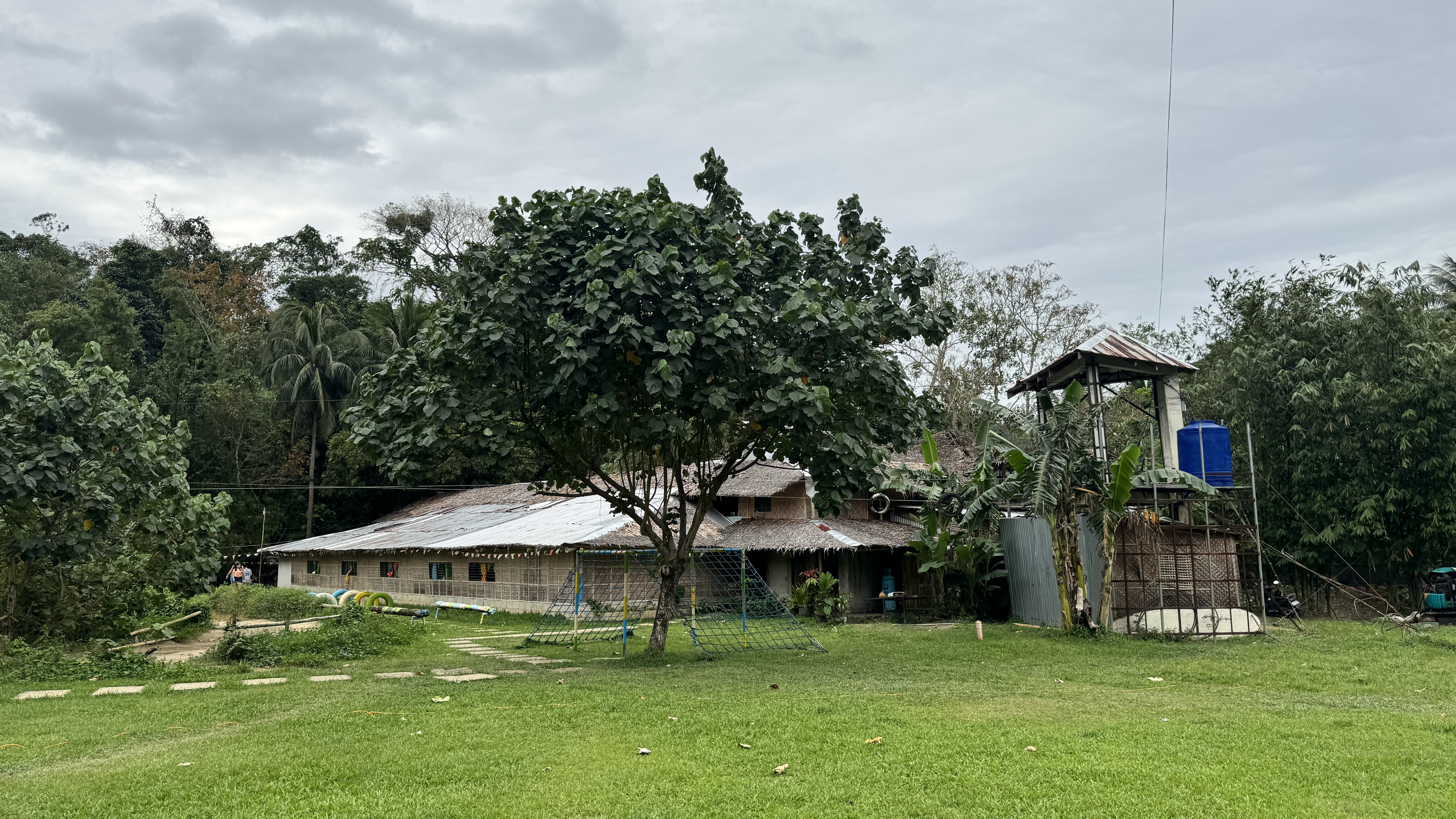 A green backyard with a small rural house made of bamboo and tin, surrounded by trees, a water tank tower, and a playground area under a cloudy sky.