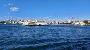 A white ferry with yellow masts is sailing on a blue body of water, with a coast lined with buildings and a large Turkish flag in the background. 