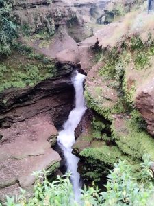 

A  devid fall waterfall cascading through narrow rock formations, surrounded by lush greenery and moss-covered rocks. The water flows down into a crevice, creating a scenic natural view.