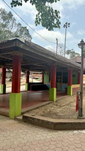 An open-air structure with a tiled roof supported by red pillars with green bases, surrounded by trees and a partly cloudy sky. The building features colorful walls in green, red, and blue, with decorative elements and a barred window. The foreground includes a paved area with interlocking tiles, a streetlamp, and a floodlight pole, with other trad
