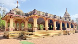 A vibrant Indian temple with a mustard-yellow facade, adorned with intricate red and orange designs, featuring multiple domed spires topped with flags. The temple includes an open pavilion with elaborately carved pillars, accessible by steps, set against a backdrop of lush greenery and a brick-paved courtyard.