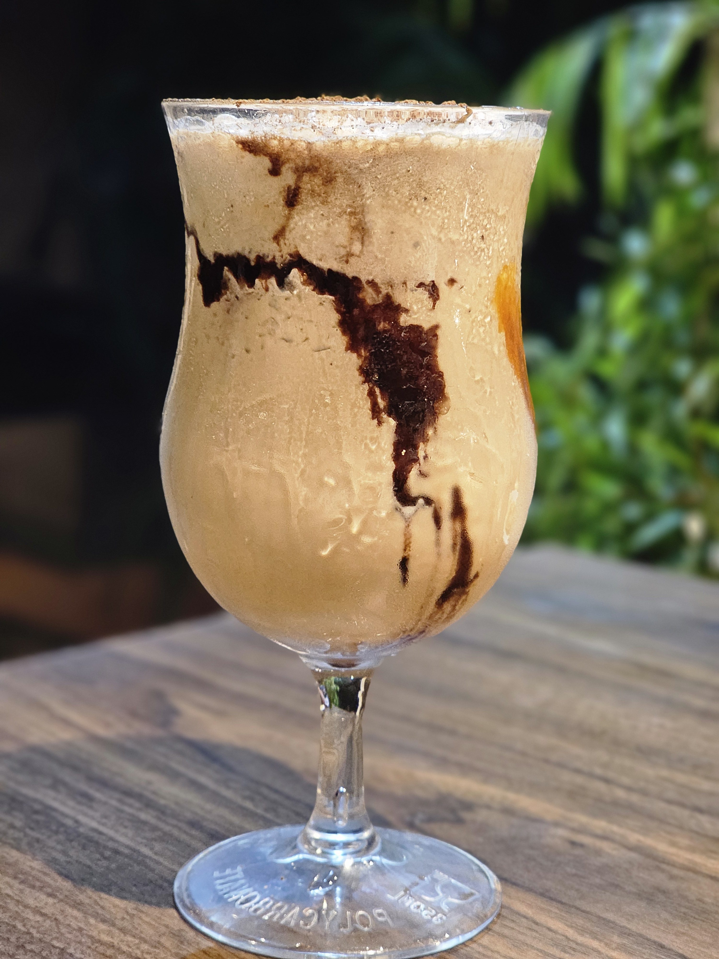 A glass of Café Frío, a cold coffee beverage, with chocolate sauce drizzled inside. The drink is served in a stemmed glass, and condensation is visible on the outside, indicating it is chilled, and the background features out-of-focus greenery. Captured from Kozhikode, Kerala.