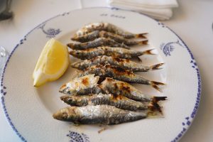 A plate of grilled sardines lined up neatly, with a slice of lemon placed next to them. The fish are browned and crispy, showcasing a smoky texture, and are arranged on a decorative plate featuring a blue and white design.
