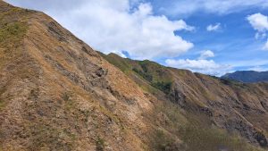 A scenic view of dry, rugged hills with patches of green vegetation under a partly cloudy blue sky.