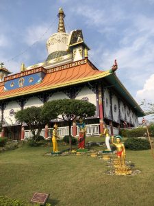 A colorful Buddhist temple with ornate architectural details, featuring a large stupa and statues of figures in traditional attire on a neatly maintained grassy area. The sky is clear and sunny.